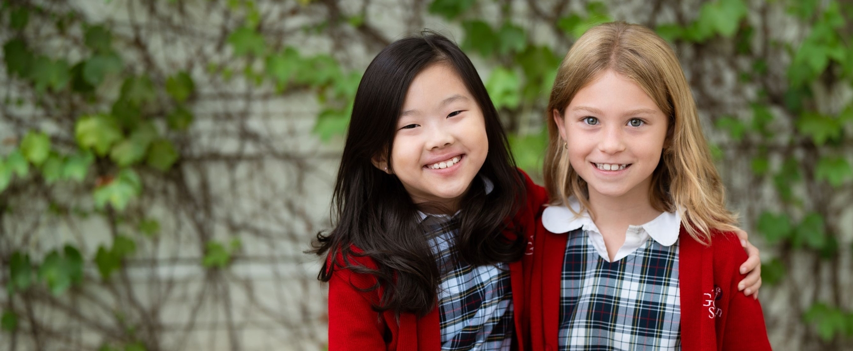 Two smiling students in matching plaid uniforms and red cardigans sit side-by-side.