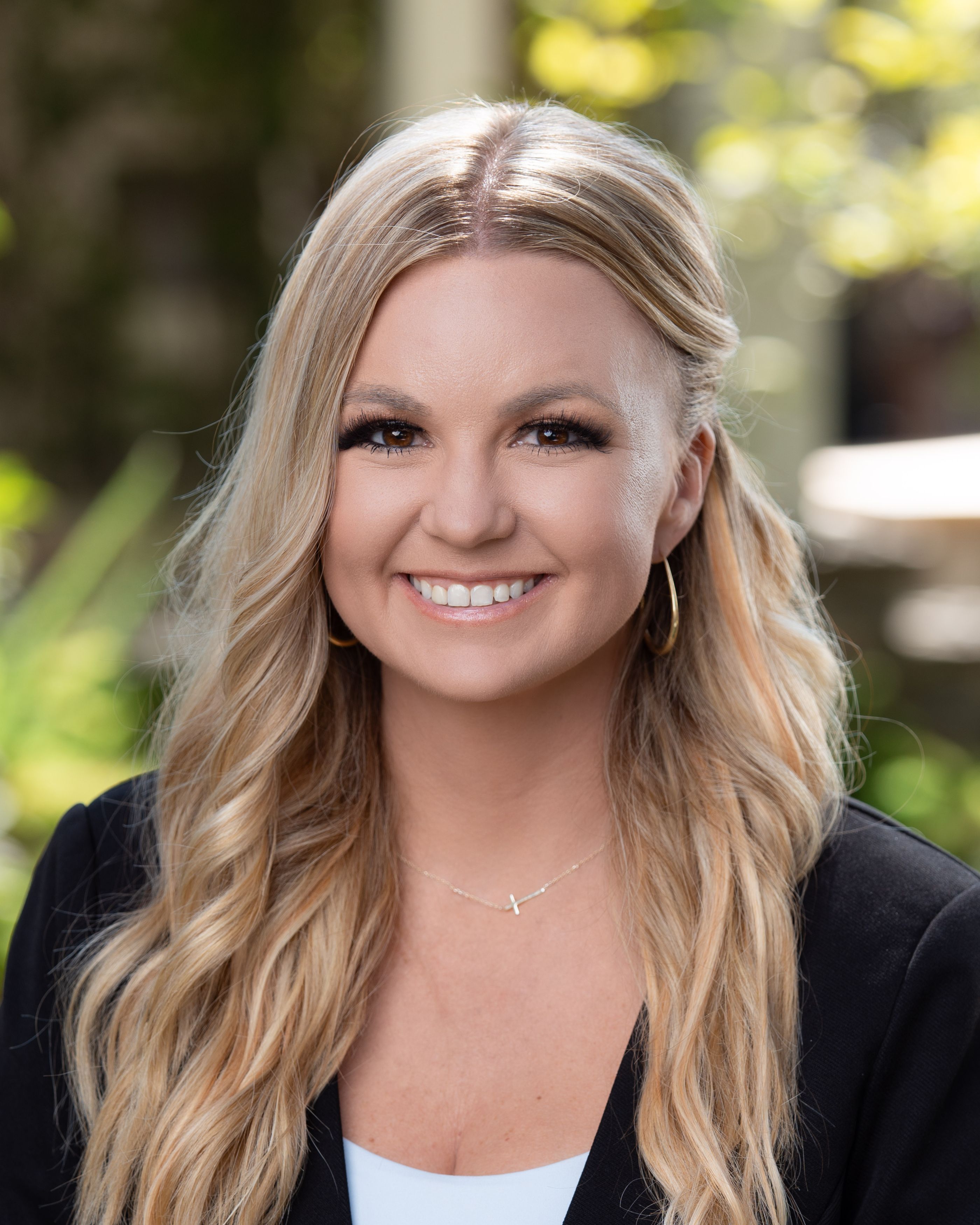 Smiling woman with long blonde hair in a black blazer, wearing hoop earrings and a small cross necklace outdoors.
