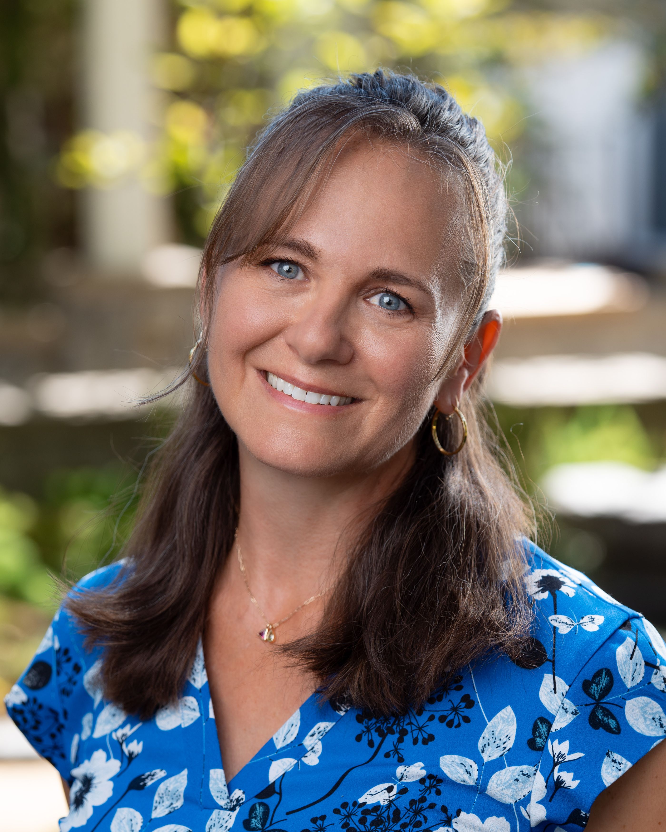 Smiling woman with long brown hair in a blue floral blouse poses outdoors with blurred greenery in the background.