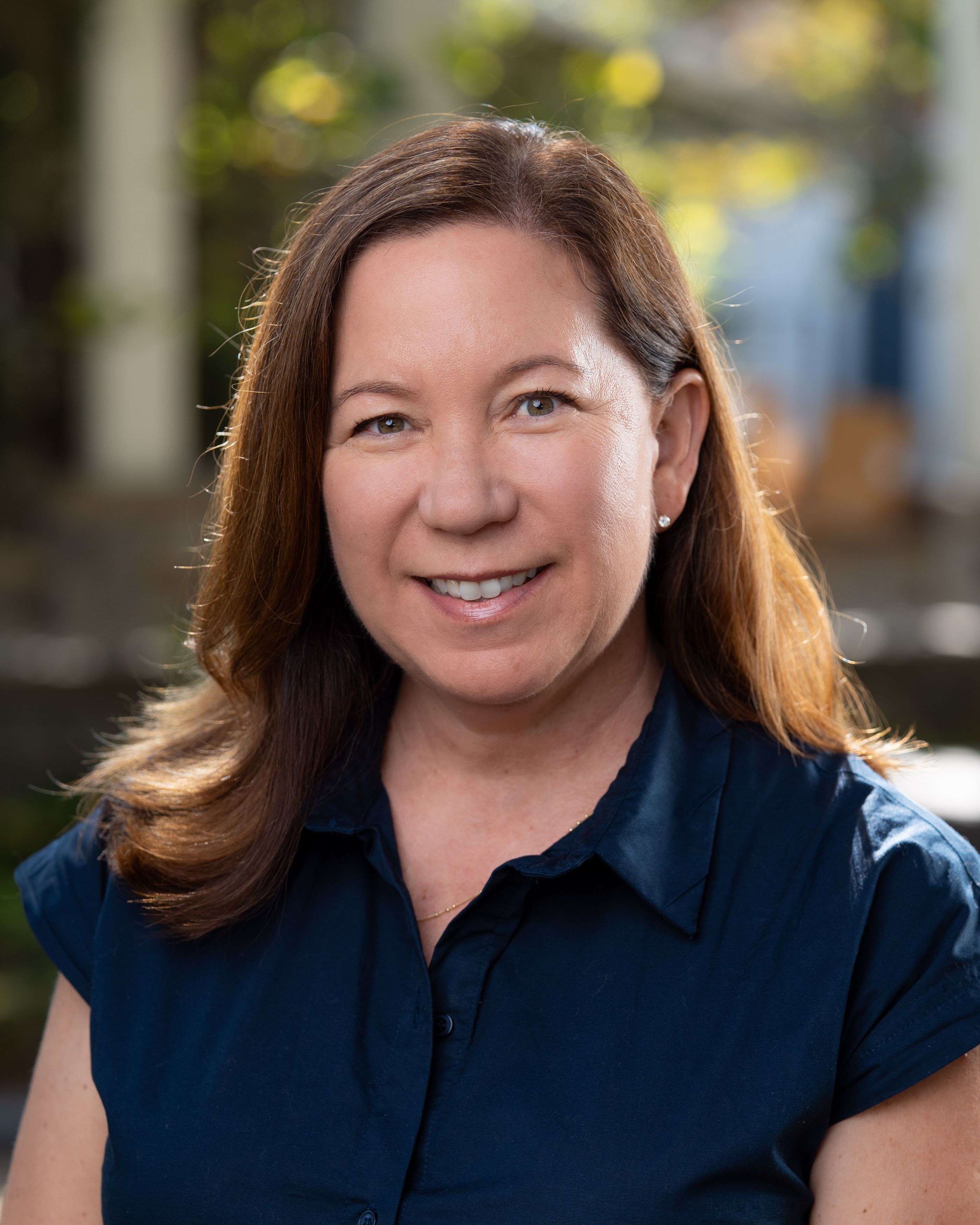 Woman smiling outdoors in soft daylight, wearing a navy collared shirt with blurred greenery in the background.
