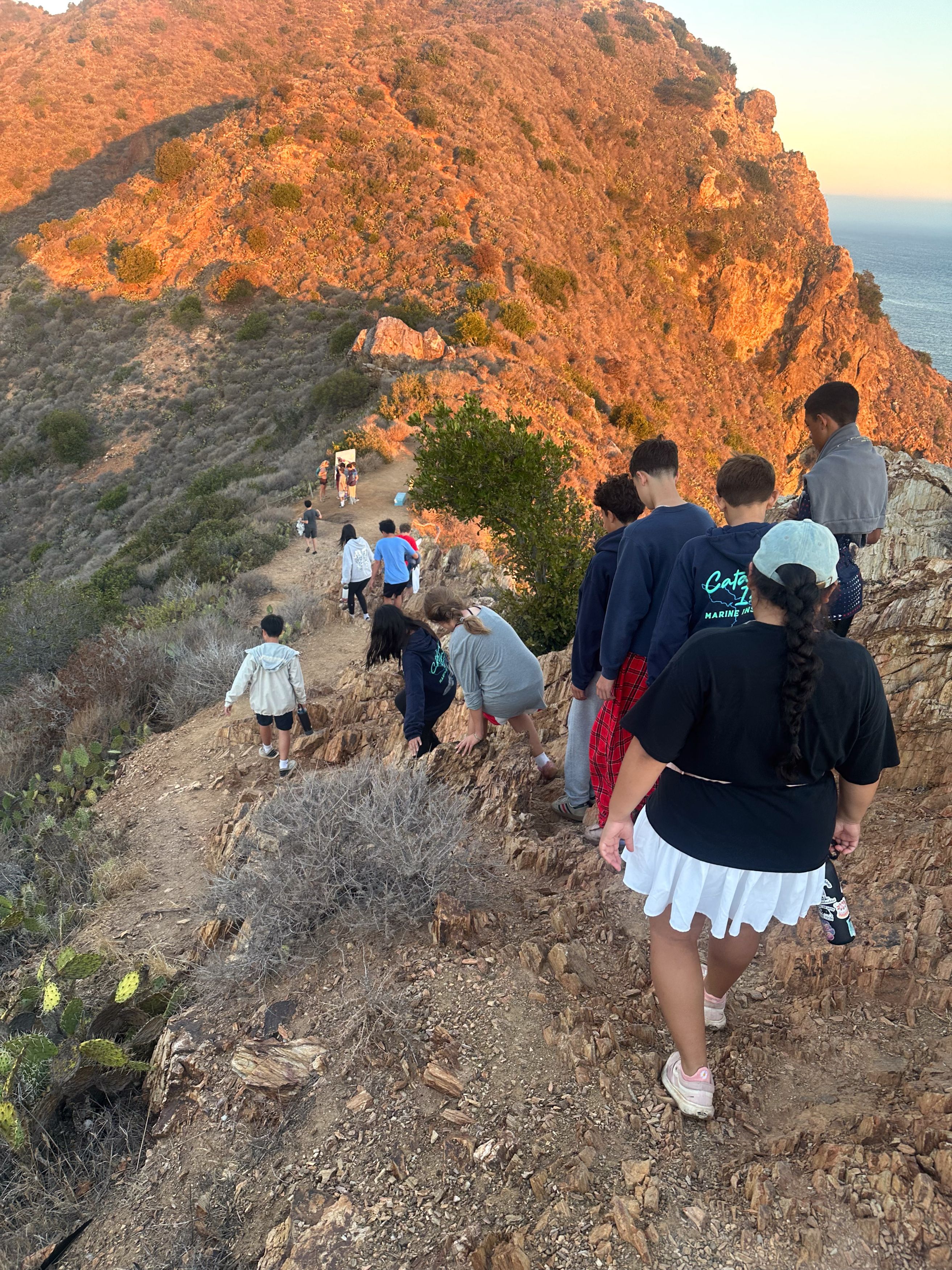 A group of students hikes down a rocky mountain trail toward the ocean at sunset.