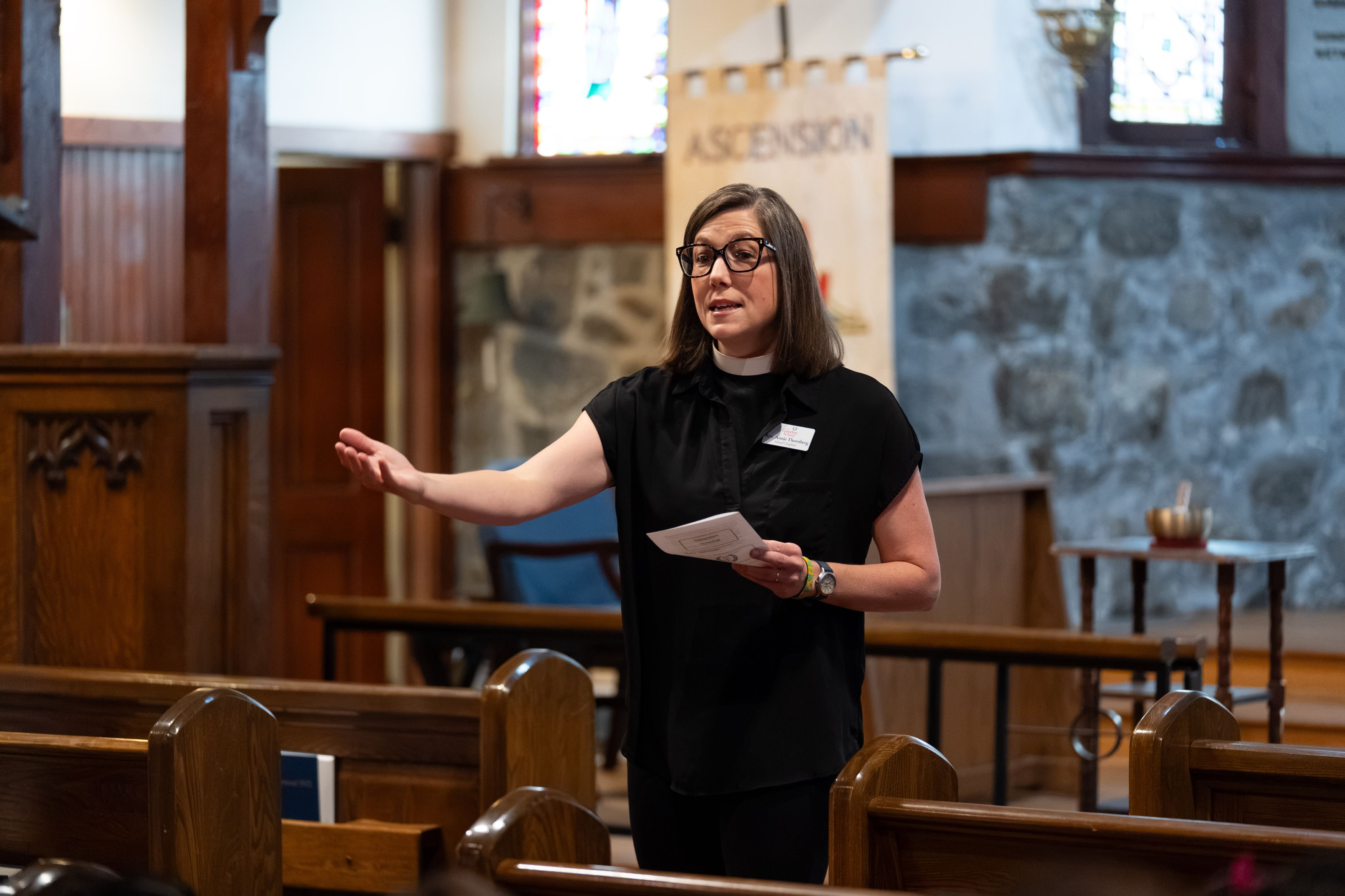 A woman wearing a clerical collar speaks from the aisle of a church, gesturing with one hand and holding notes in the other.