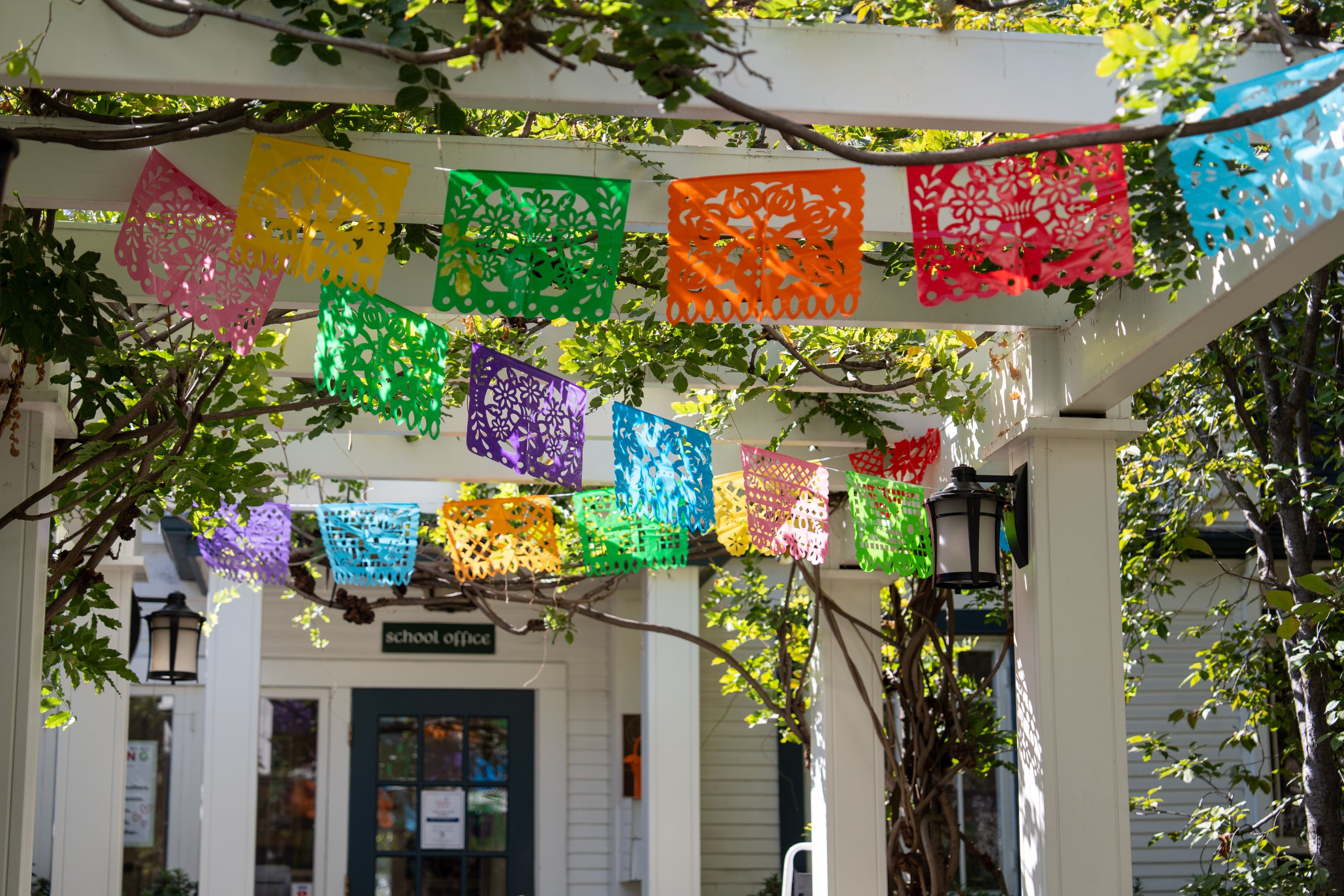 Colorful papel picado banners hang from a white pergola outside a building with a sign reading School office.