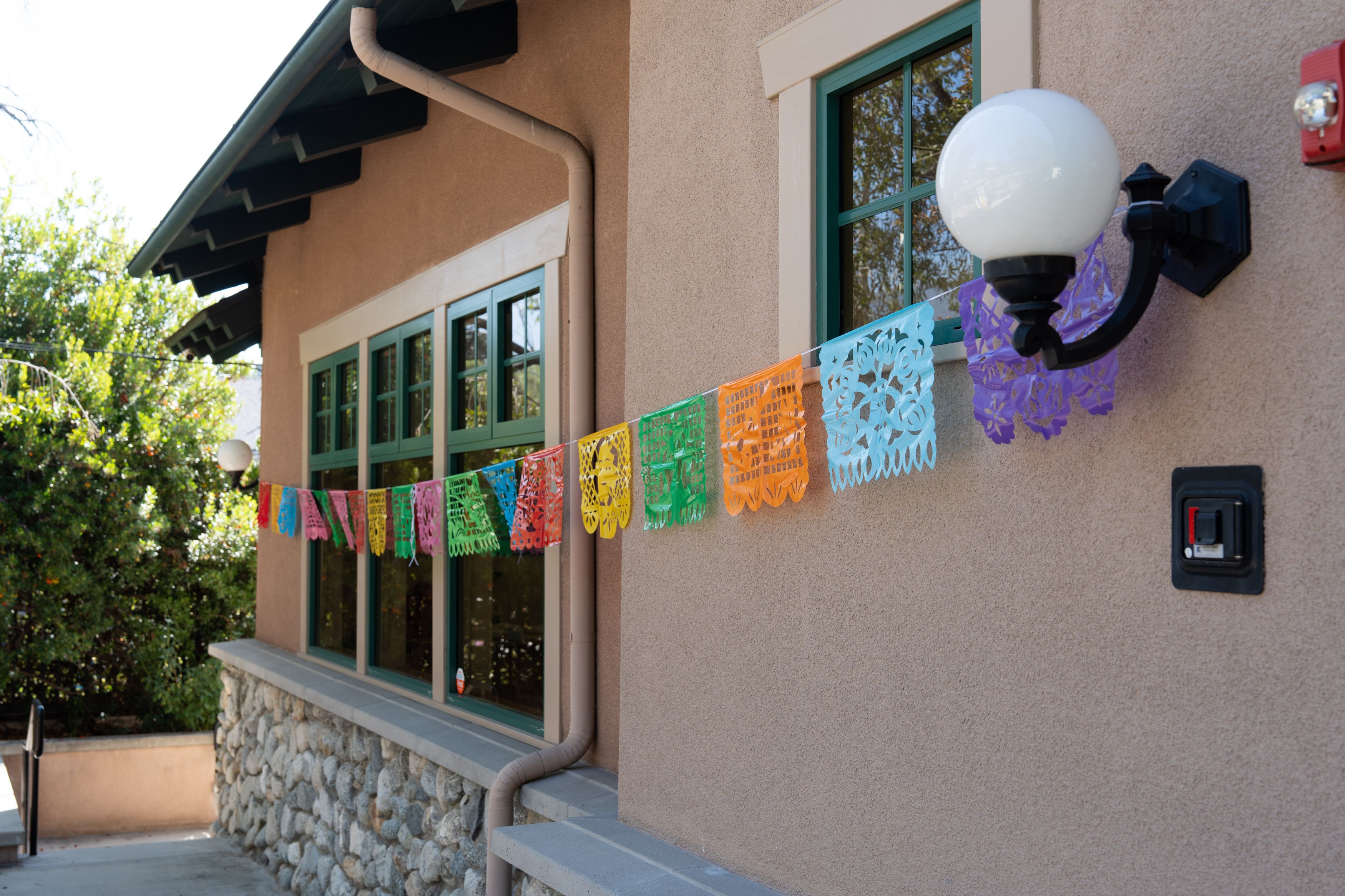 Colorful papel picado banners hang across the exterior of a stucco building with green-trimmed windows.