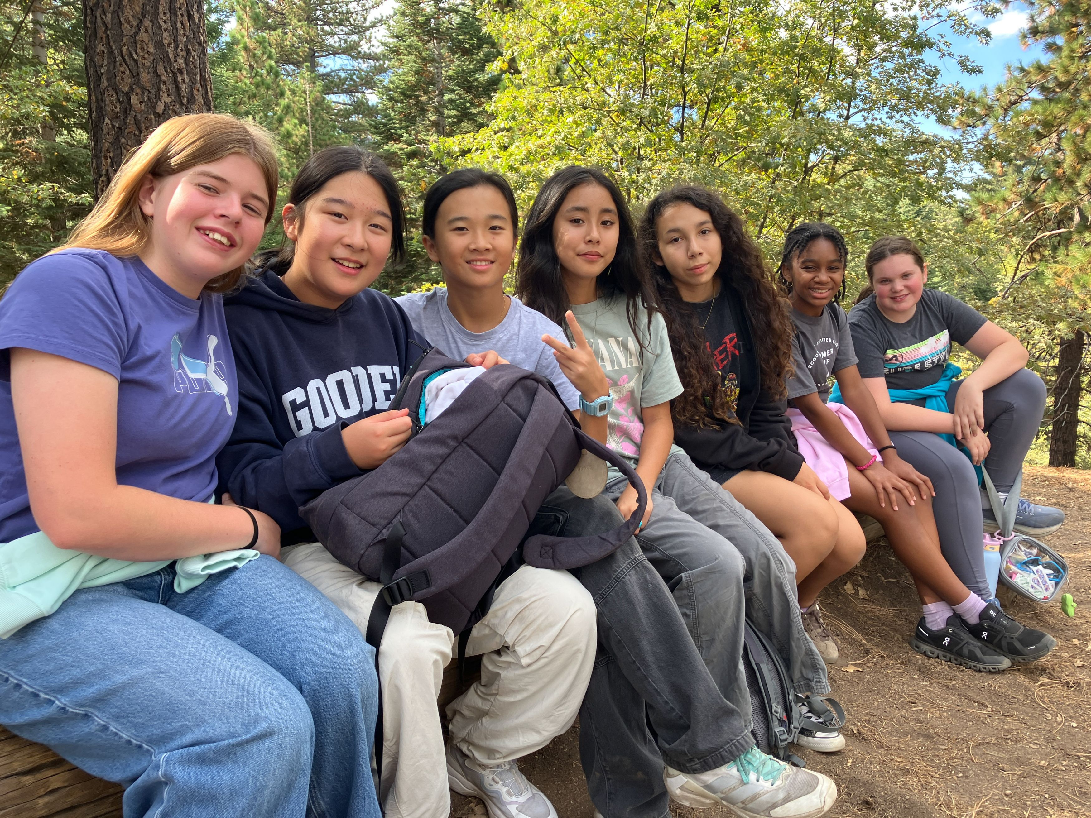 Seven students from The Gooden School sit closely together on a log outdoors, smiling at the camera.