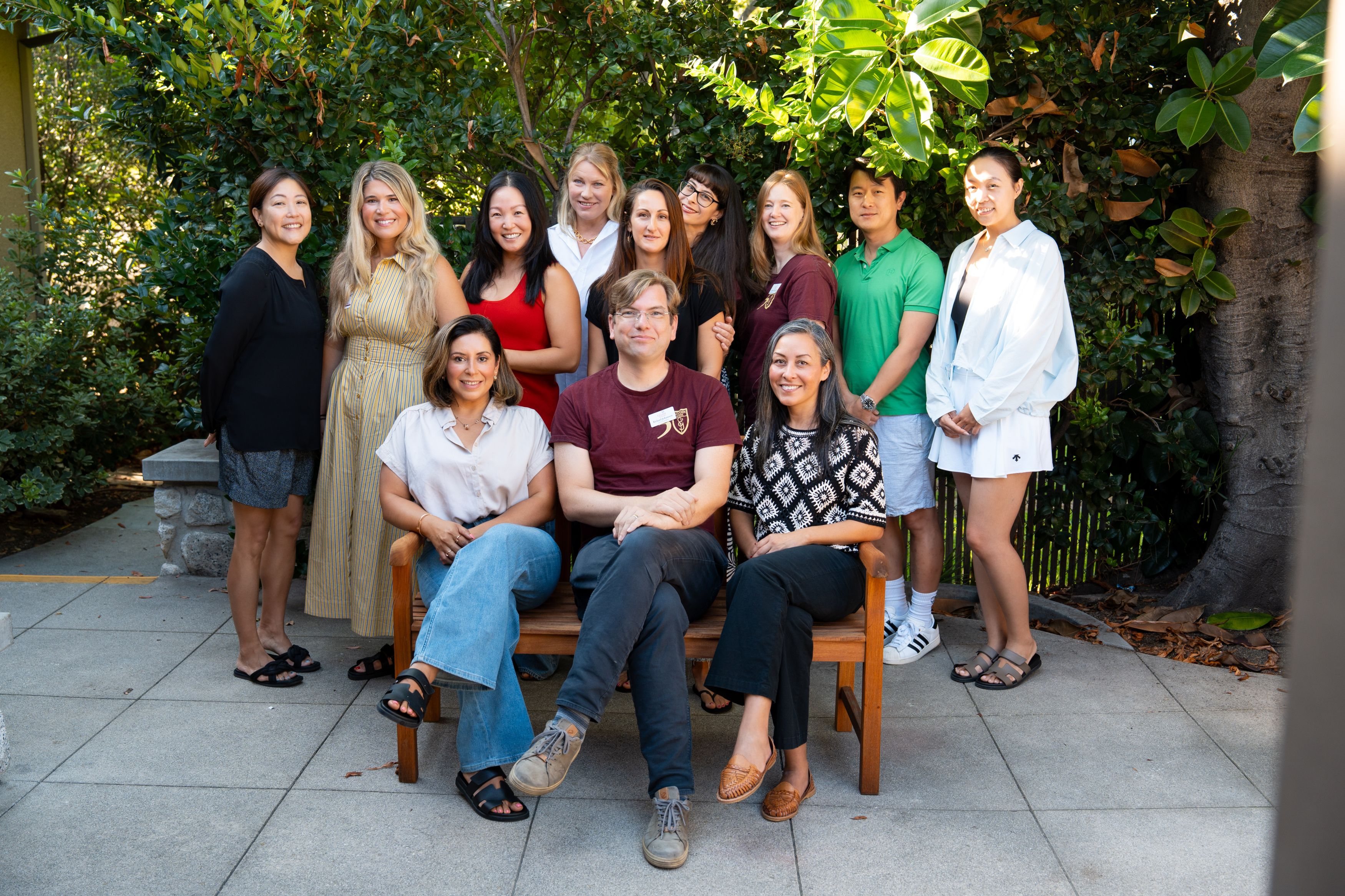 Twelve adults pose outdoors in two rows, with three seated on a wooden bench and ten standing behind amid lush greenery.